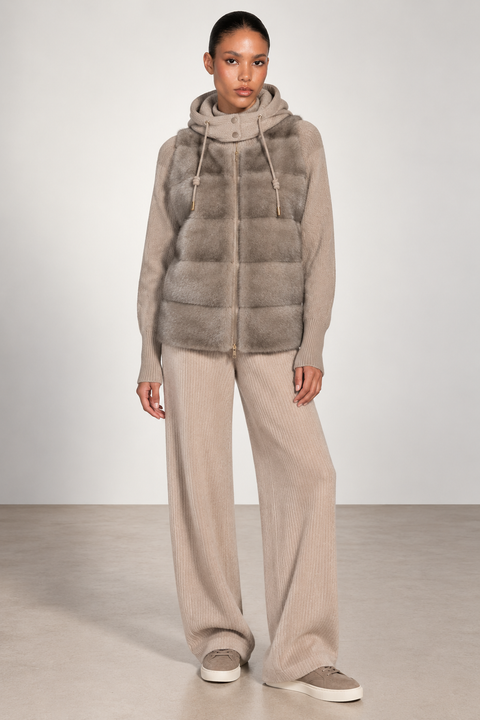 A model stands in a studio wearing a taupe faux-fur hooded puffer vest over a matching taupe knit hoodie and wide-leg knit pants, with taupe slip-on shoes.