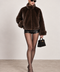 A model wearing a dark brown faux fur jacket, black leather shorts, sheer black tights, and black pointed-toe heels, holding a small black handbag against a neutral studio backdrop.